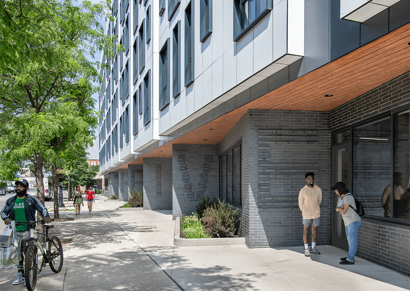 People interacting near a building with large windows and a sleek exterior. A cyclist is seen on the left side of the image.