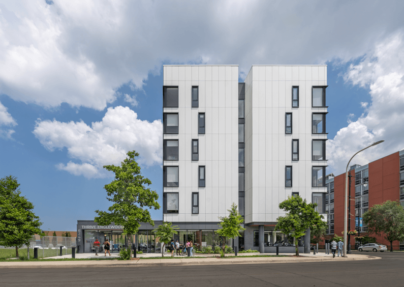 New apartment building with large windows, located in Englewood. Green landscaping with trees and shrubs in the foreground. People visible near the entrance.