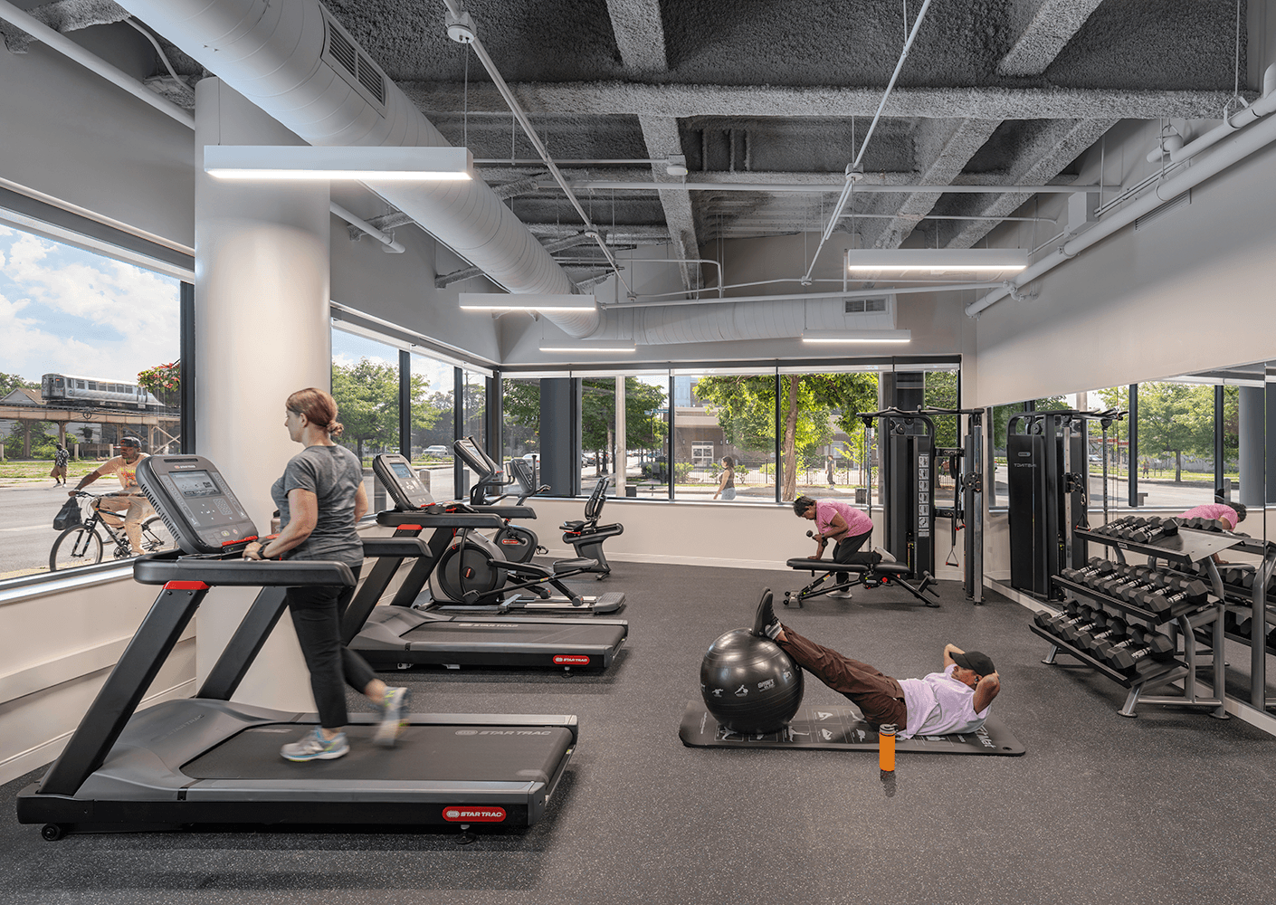 Two individuals workout in a gym with large windows, featuring treadmills, weights, and exercise equipment in a bright space.