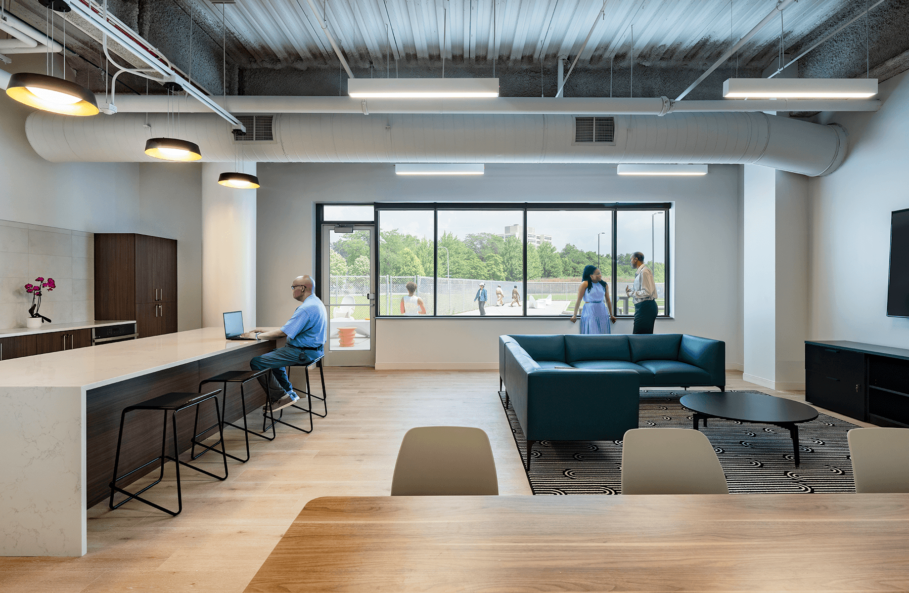 A stylish communal workspace featuring a kitchen area, seating, and large windows. Individuals work and interact in a vibrant atmosphere.