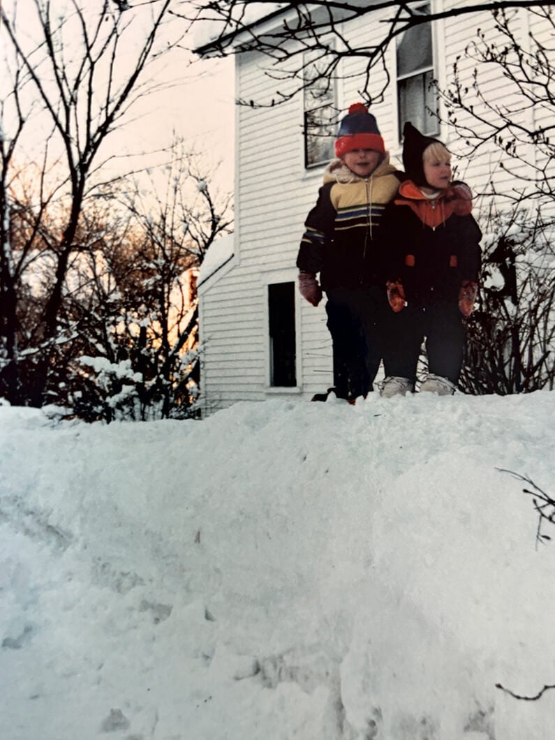 Two children in winter clothing stand on a snow mound beside a white house, with trees and a sunset in the background.