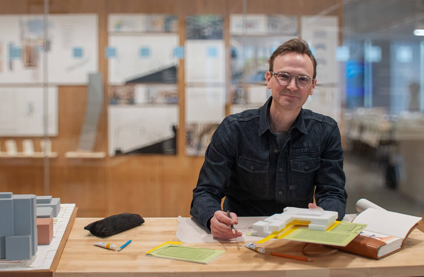 Architect at a workspace, drawing plans and modeling structures. Papers, tools, and a foam model are visible on the desk.