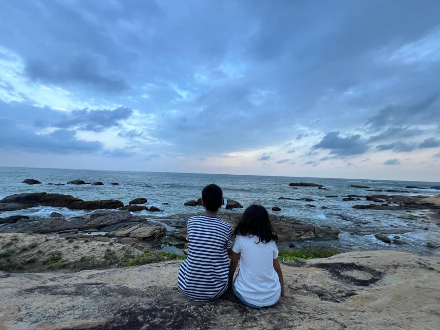 Two children sitting on a rocky shore, facing the ocean under a cloudy sky, with gentle waves lapping at the rocks.