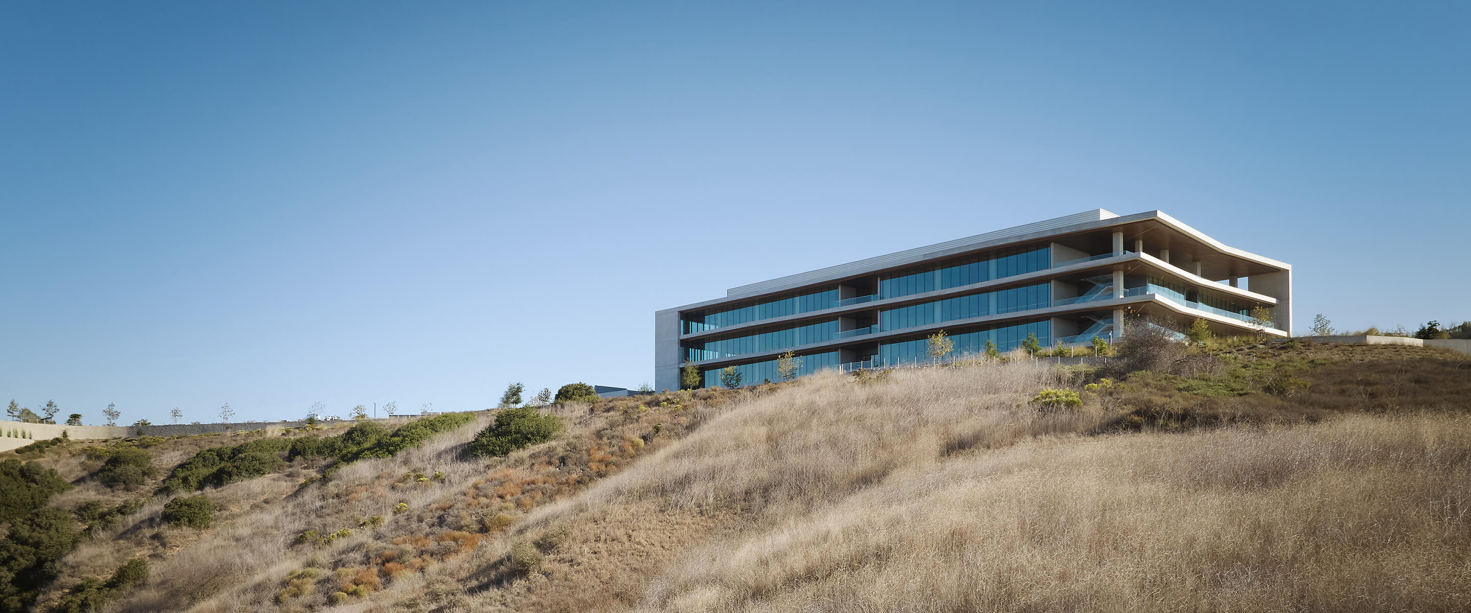 Contemporary building on a hillside, featuring large glass windows and multiple levels, surrounded by dry grass and sparse vegetation under a clear sky.