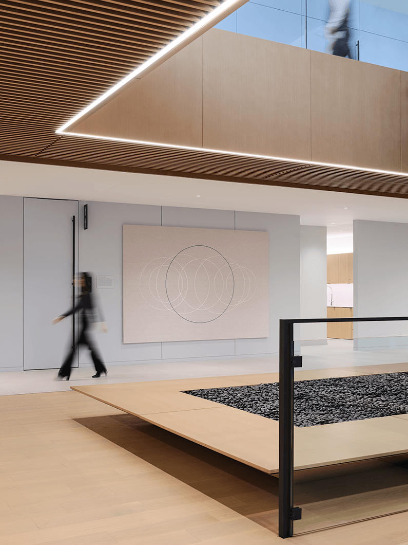 Interior view of a spacious lobby featuring wooden ceiling panels, a stone-filled area, and a large wall art piece, with a person walking by.