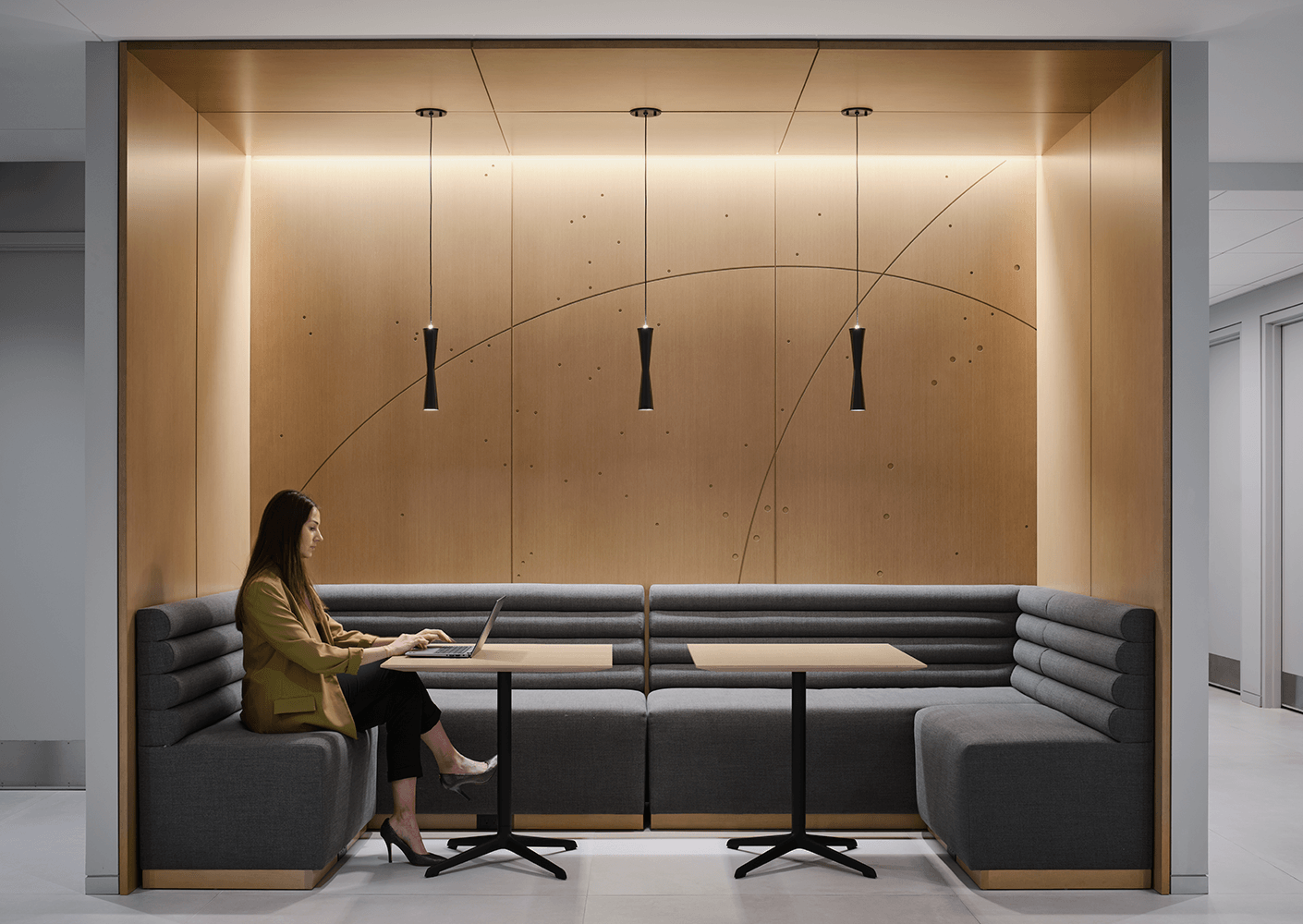 A woman working on a laptop sits in a cozy seating area with wooden and grey accents, surrounded by soft lighting and minimalist decor.