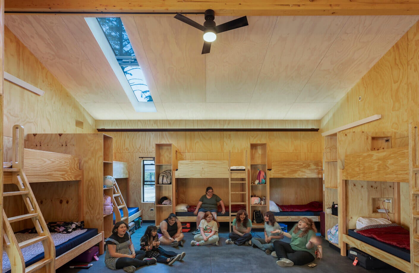 Group of people sitting on the floor of a room with wooden bunk beds, large windows, and a ceiling fan. Natural light illuminates the space.