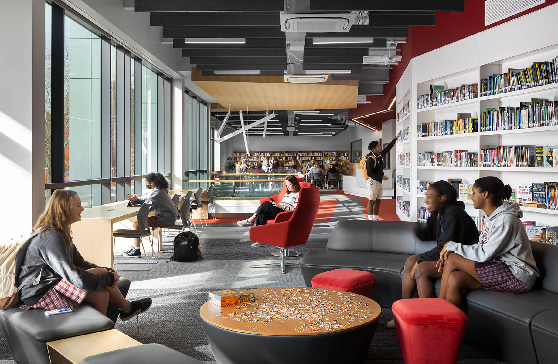 Students engage in study and leisure activities in a library with large windows, comfortable seating, and bookshelves in the background.