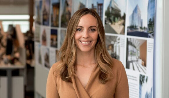 A woman with long hair smiles in front of architectural design boards in an office setting, wearing a tan sweater.