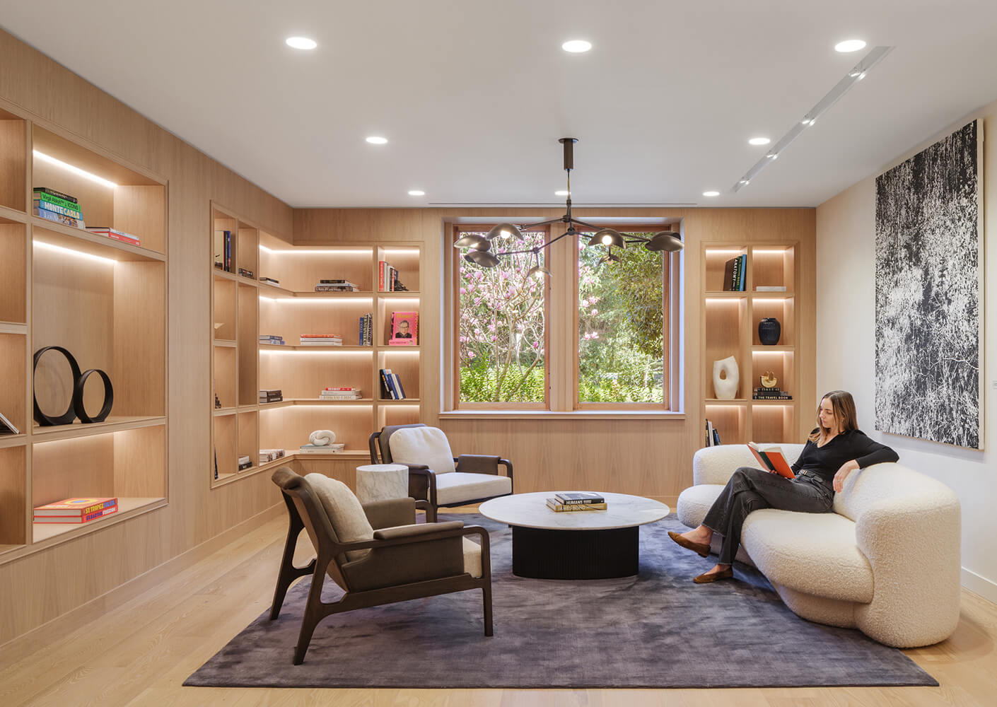 Cozy reading area with soft seating, a marble coffee table, and wooden shelves showcasing books and decorative items. Natural light filters in from windows.