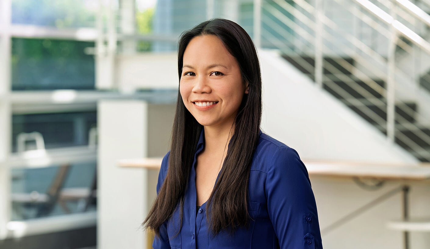 Professional woman with long black hair, wearing a blue shirt, smiling in an office setting with a staircase and natural light in the background.