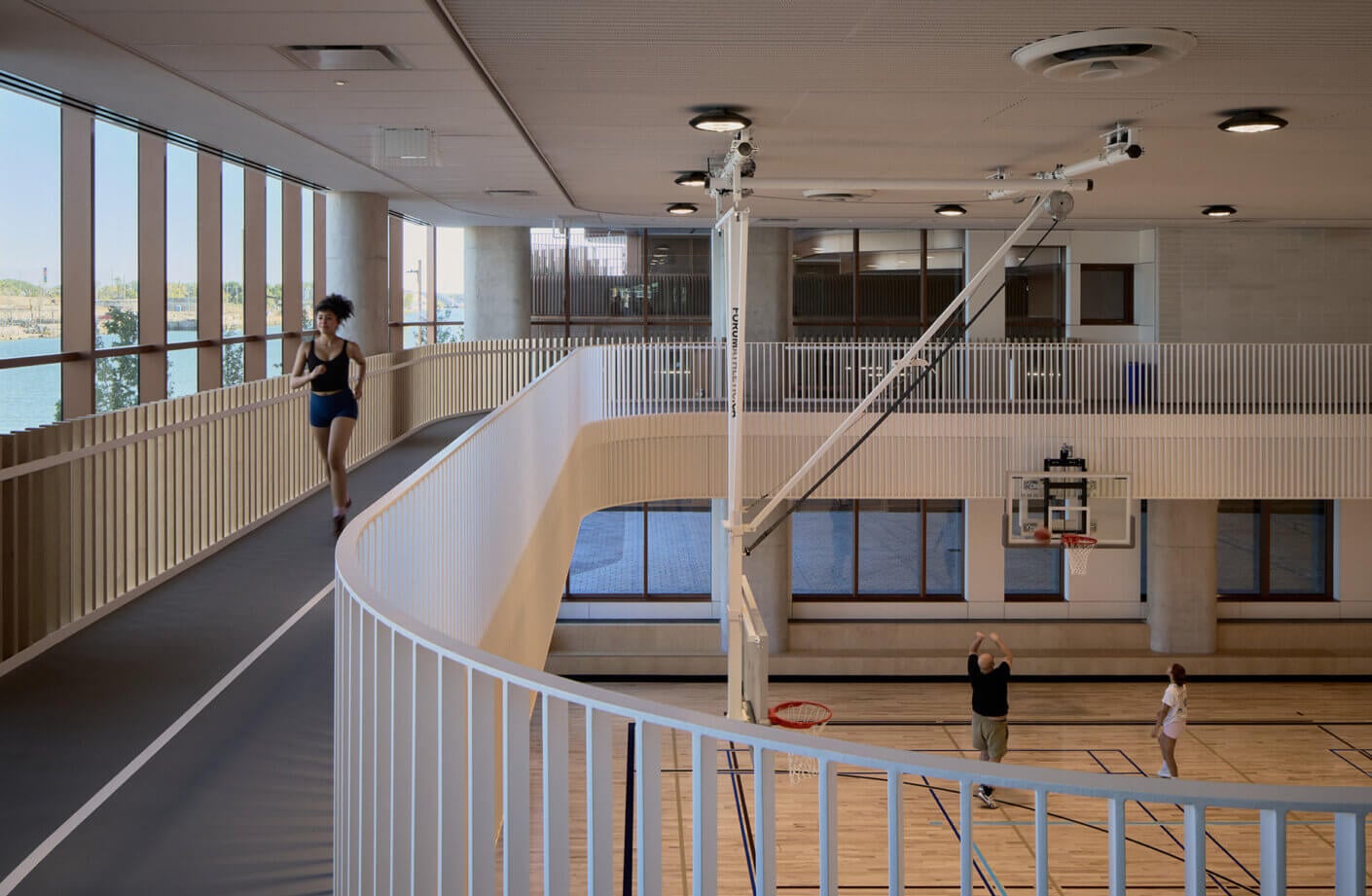 Indoor sports facility featuring a running track, basketball court, and large windows providing natural light. People engaging in activities.