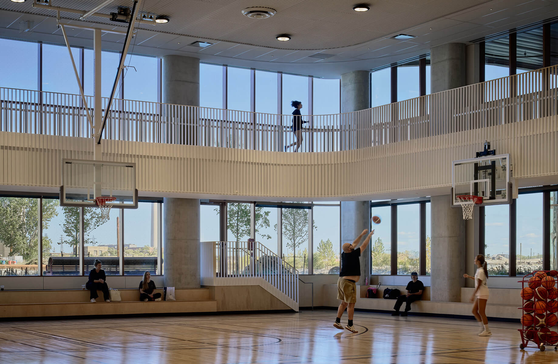 Person shooting a basketball in a gym with large windows. Two spectators sit on benches, while another person walks on a balcony above.