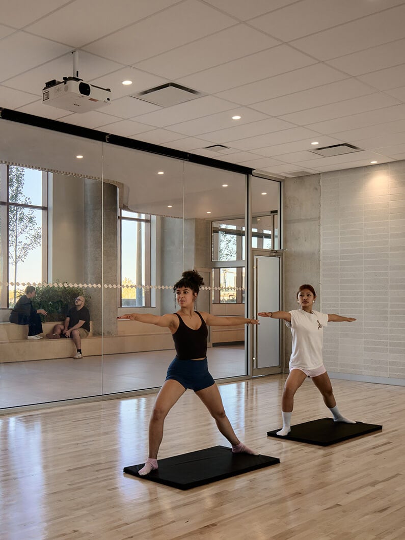 Two individuals practice yoga poses in a well-lit studio with mirrors and plants. A projector is mounted on the ceiling. People chat in the background.