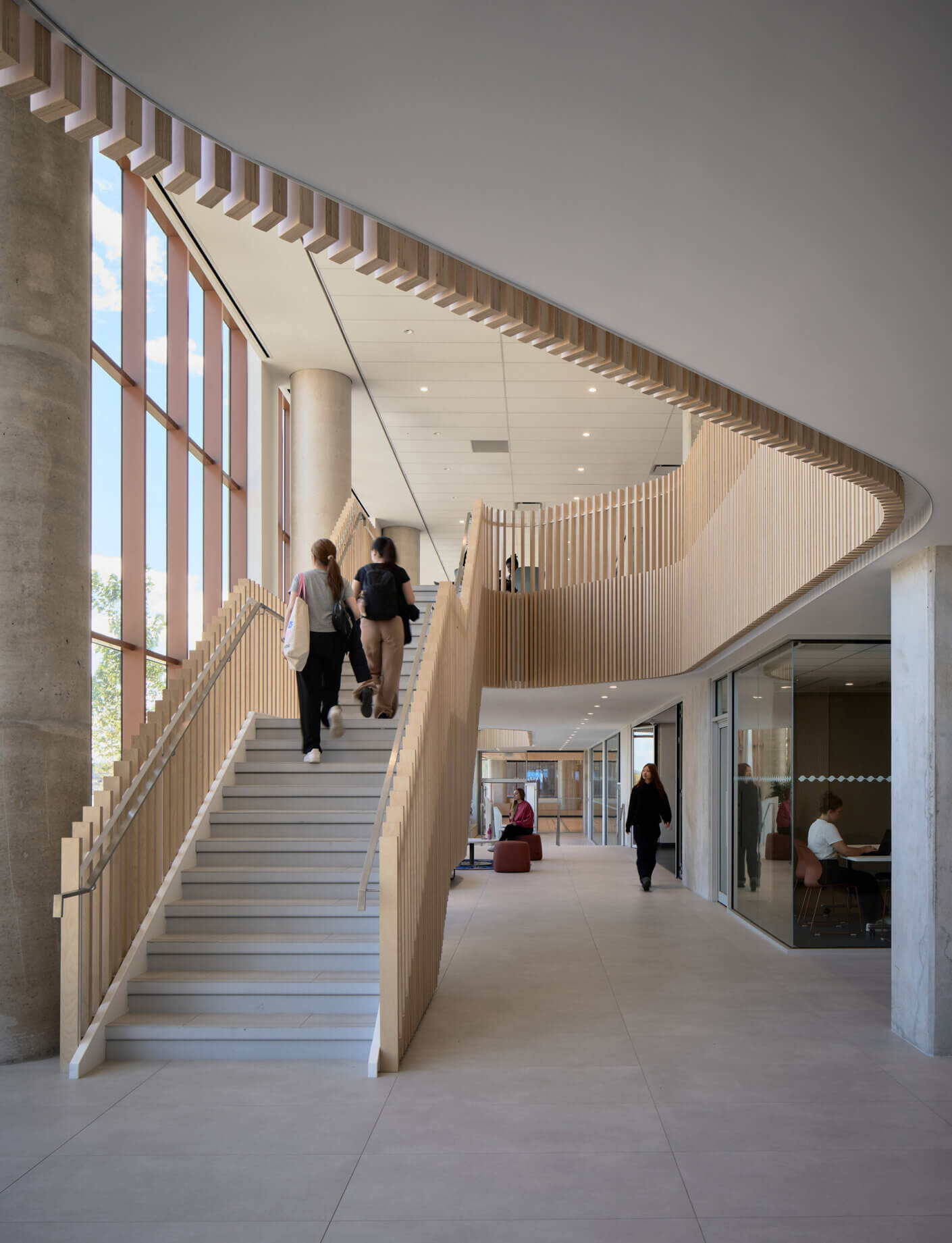 Interior view of a building featuring a staircase with wooden railing, large windows, and individuals walking through a spacious corridor.