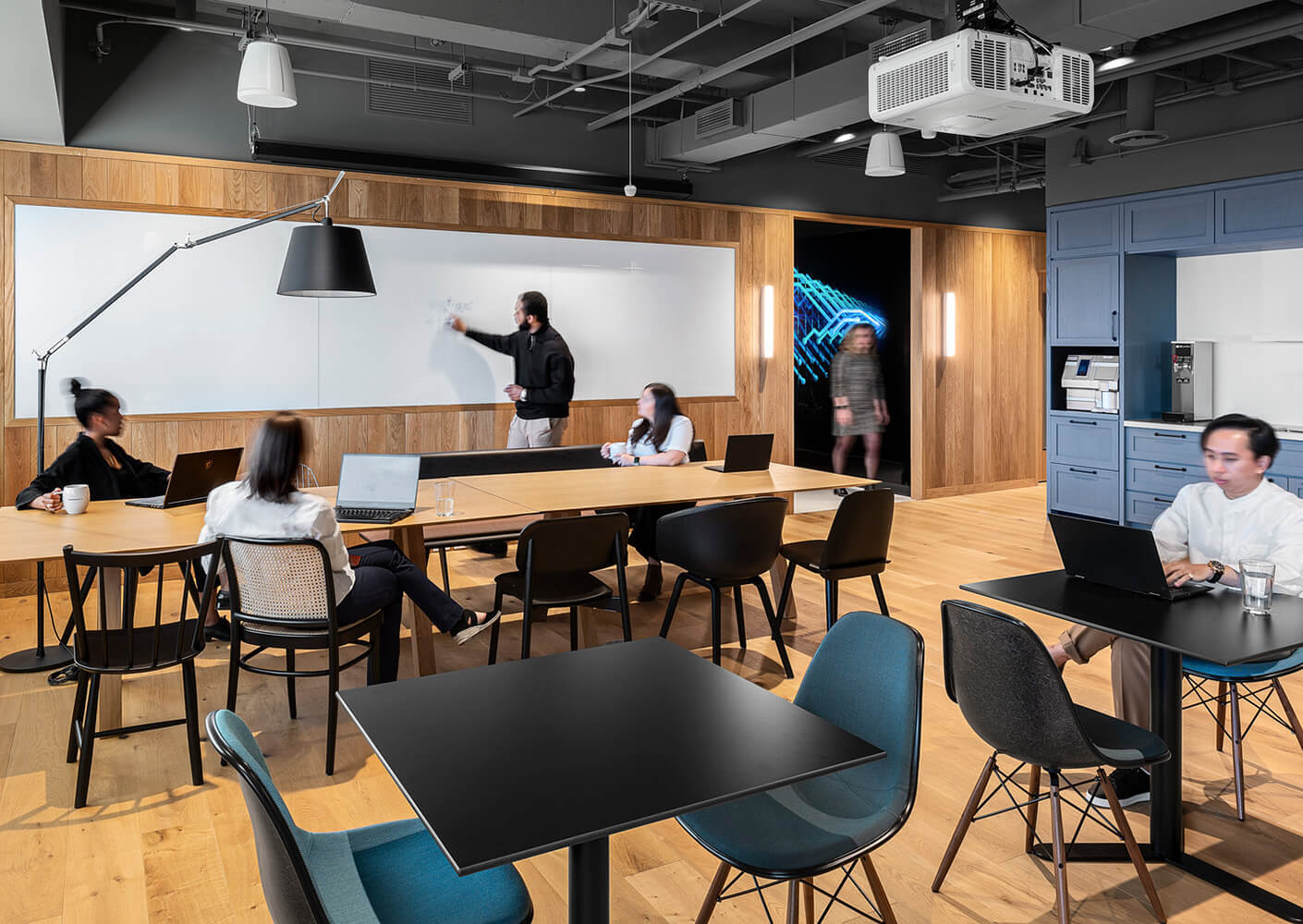 Office space with a collaborative atmosphere; people engaged in discussion, using laptops, and writing on a whiteboard. Comfortable seating and clean design.