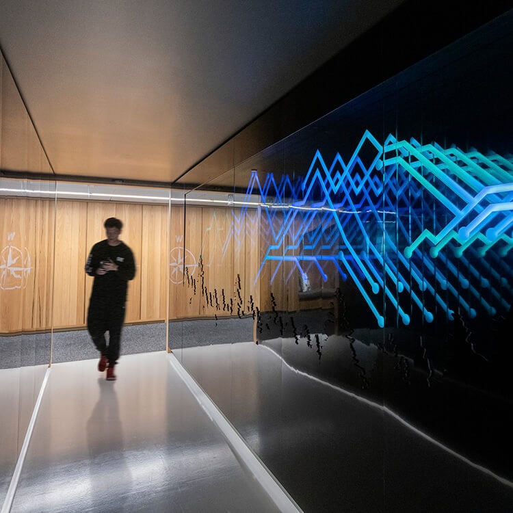 A person walking through a sleek corridor featuring a blue and green light installation against a reflective wall with wooden accents.