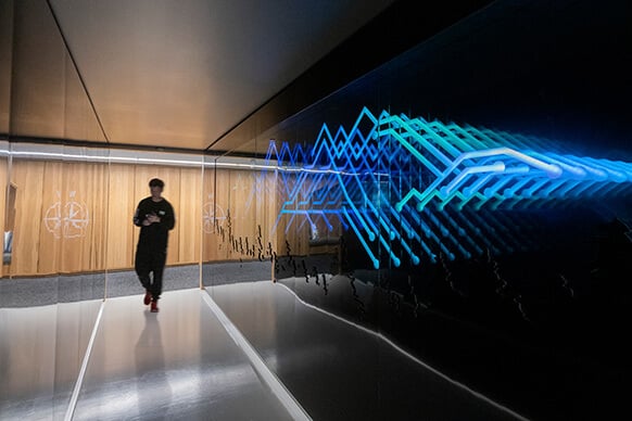 Person walking through a hallway with wood paneling, featuring a bright, abstract light display on reflective surfaces.
