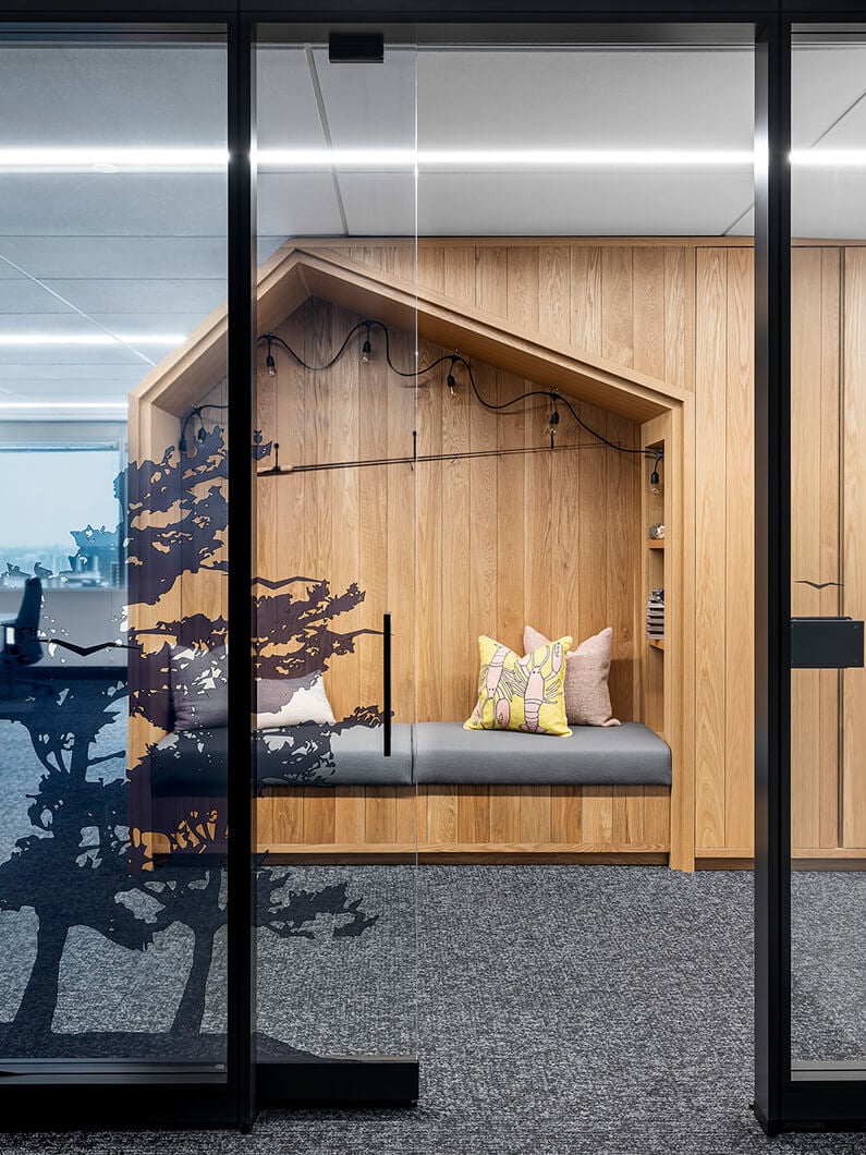 Cozy wooden nook with a bench, decorative pillows, and a glass door featuring a tree design, set in a professional office environment.