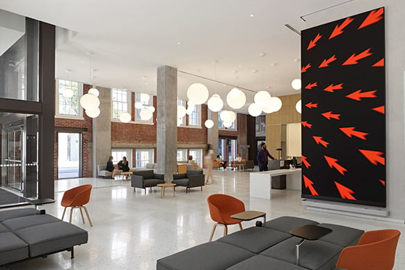 Interior view of a spacious lobby featuring seating areas, artistic wall display with arrows, pendant lights, and large windows.