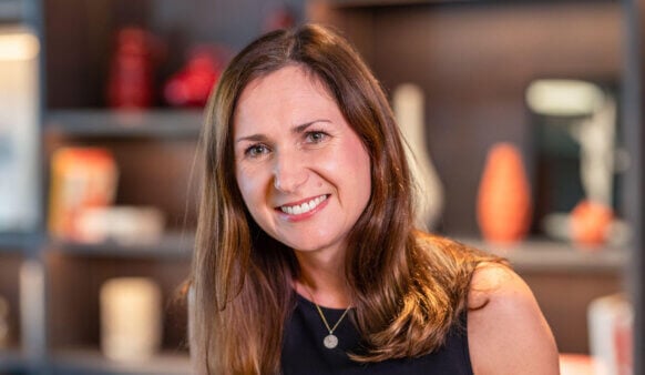 Smiling woman with long hair wearing a black top, posing in front of a shelf with decorative vases and various displayed items.