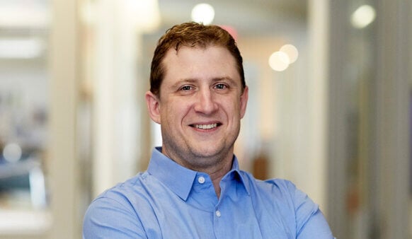 Smiling man in a light blue shirt stands with arms crossed in a well-lit hallway, featuring blurred background details of an office environment.