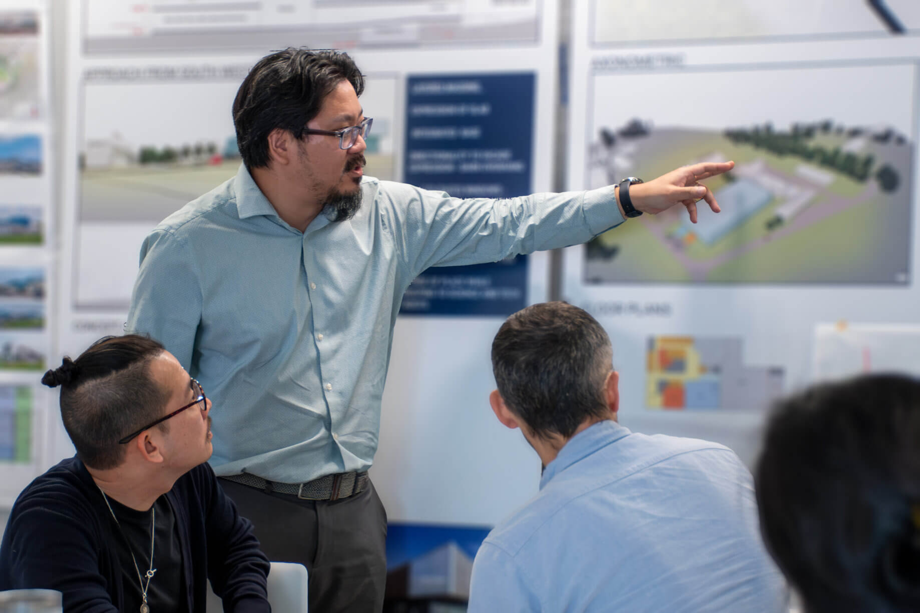 Jim Chen presents design plans to colleagues during a meeting, explaining features with gestures in front of visual displays.