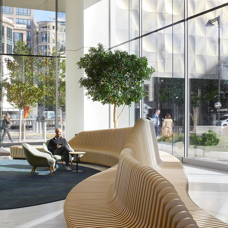 Interior of a bright lobby with large windows, featuring a curved wooden bench, a small table, and a man sitting with a smartphone. Green plants are present.