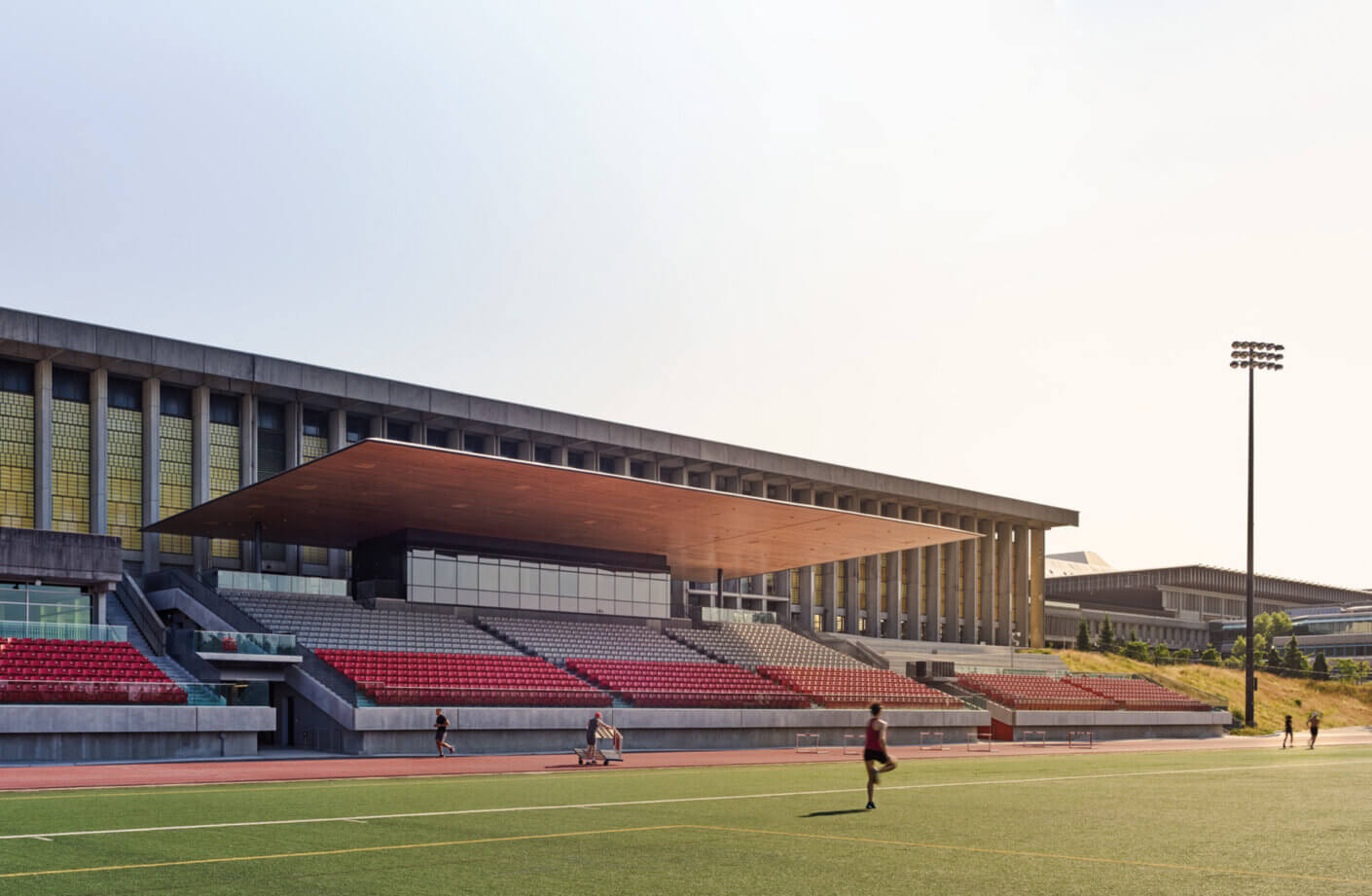 Stadium exterior with tiered seating and a wooden overhang, surrounded by a sports field and athletes in motion under clear skies.