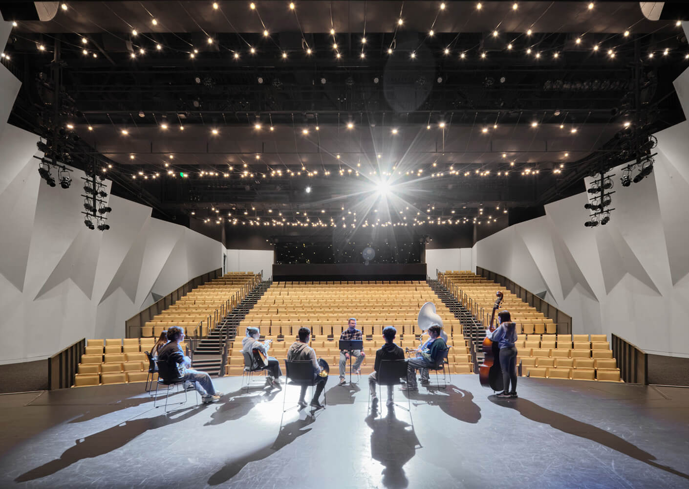 A performing arts venue with an empty audience area and musicians playing on stage, illuminated by overhead lights.