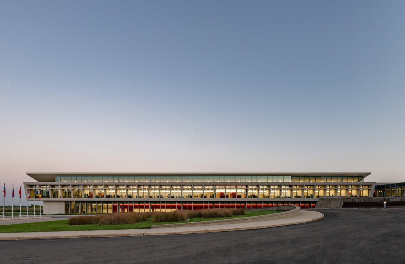 Exterior view of a sleek, glass-fronted building with landscaped grounds, showcasing flags in front and a curved access road leading to the entrance.