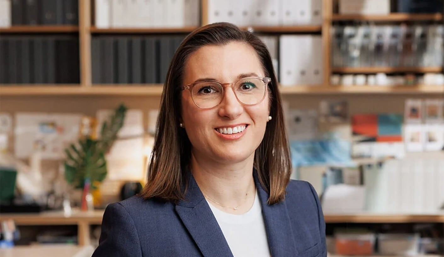Portrait of a woman smiling in an office setting, wearing glasses and a blazer, with shelves of binders and design materials in the background.