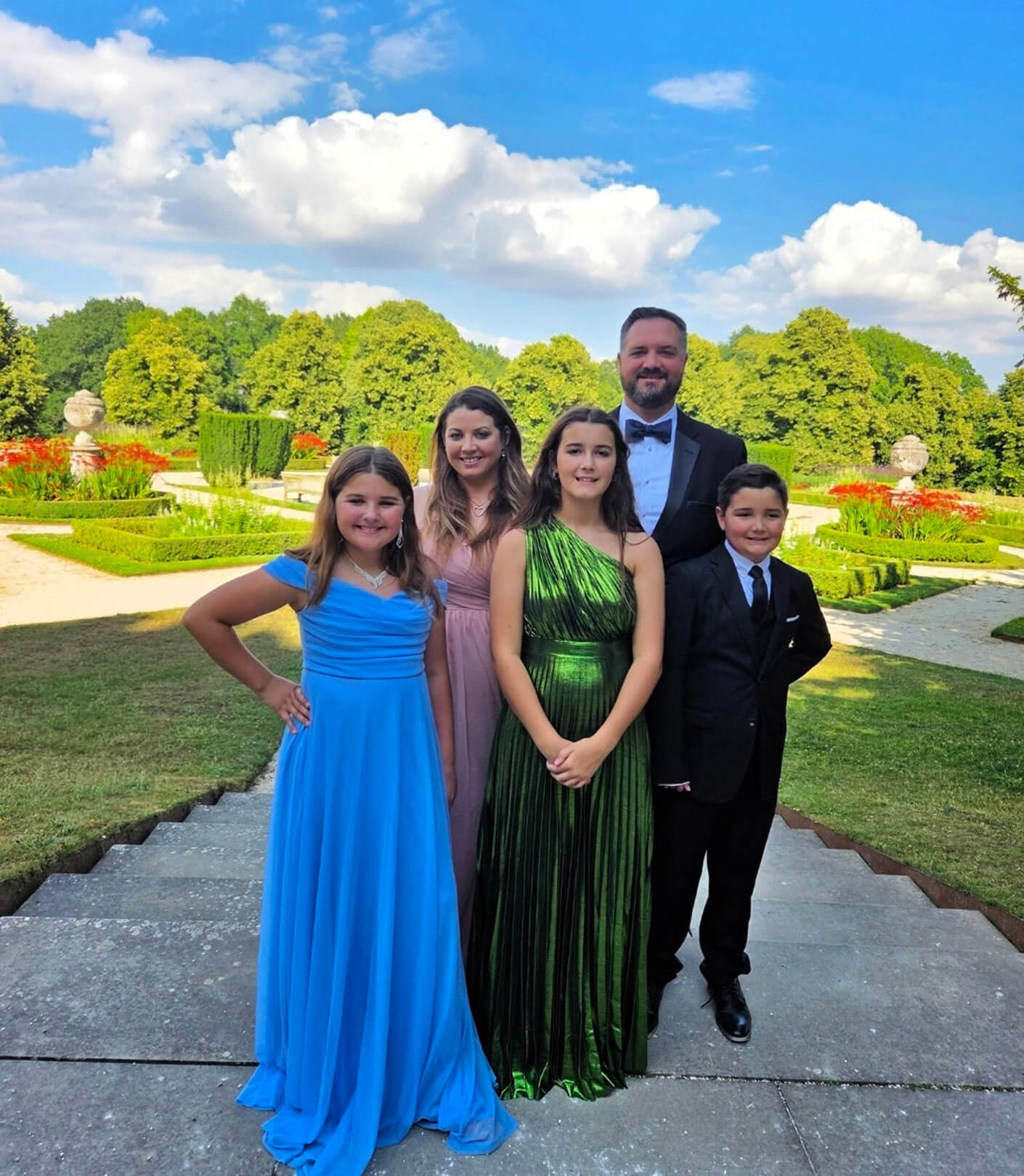 Danny and his wife with their three children, dressed in formal attire, standing in a garden with shrubs and flowers under a blue sky.