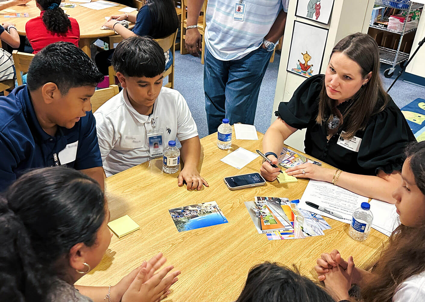 Students collaborate around a table with Lauren, reviewing pictures and taking notes in an educational setting.