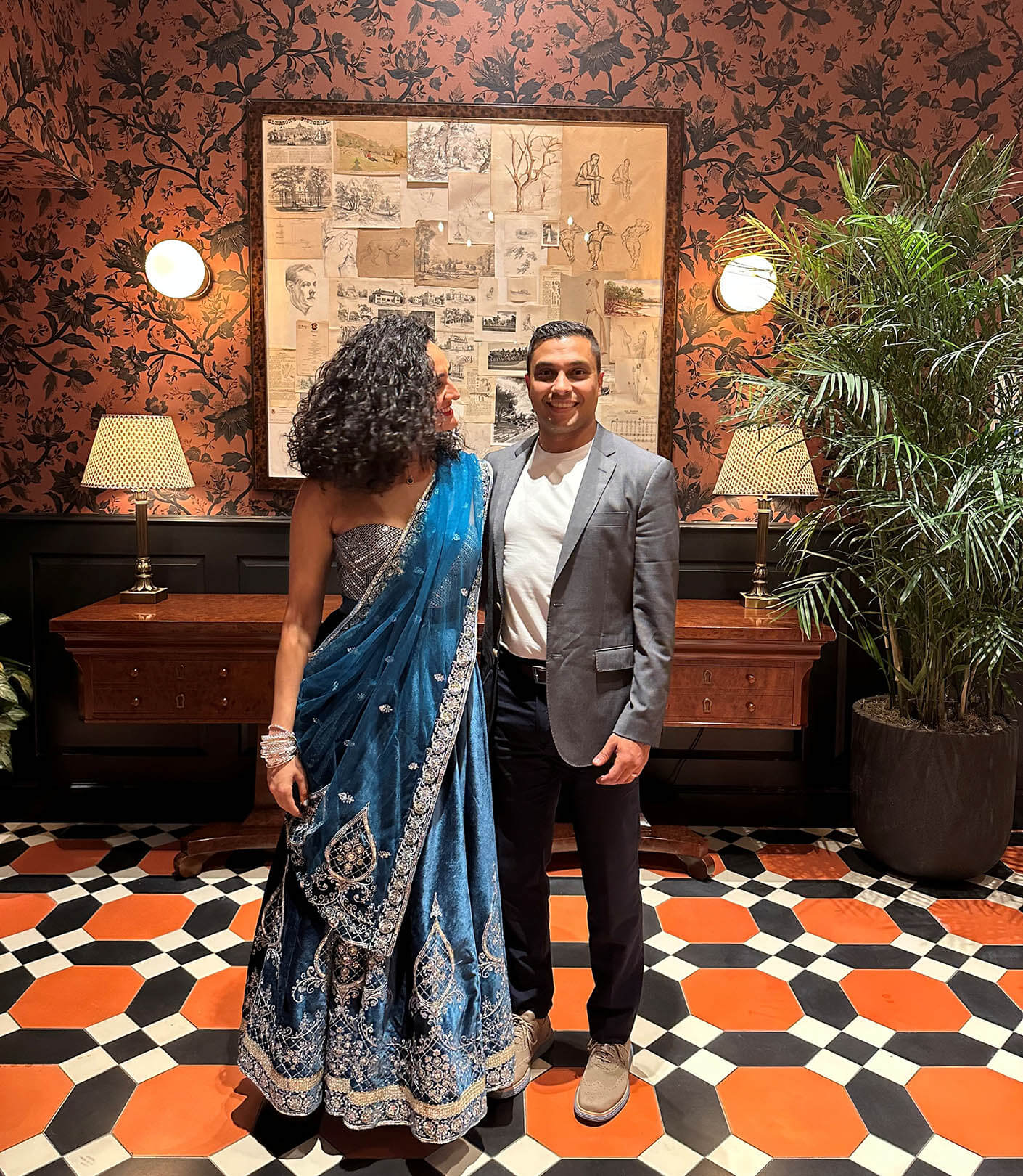 Razan poses with her husband in a decorated room with floral wallpaper; she wears a blue saree, he wears a suit. Stylish lamps and plants in view.
