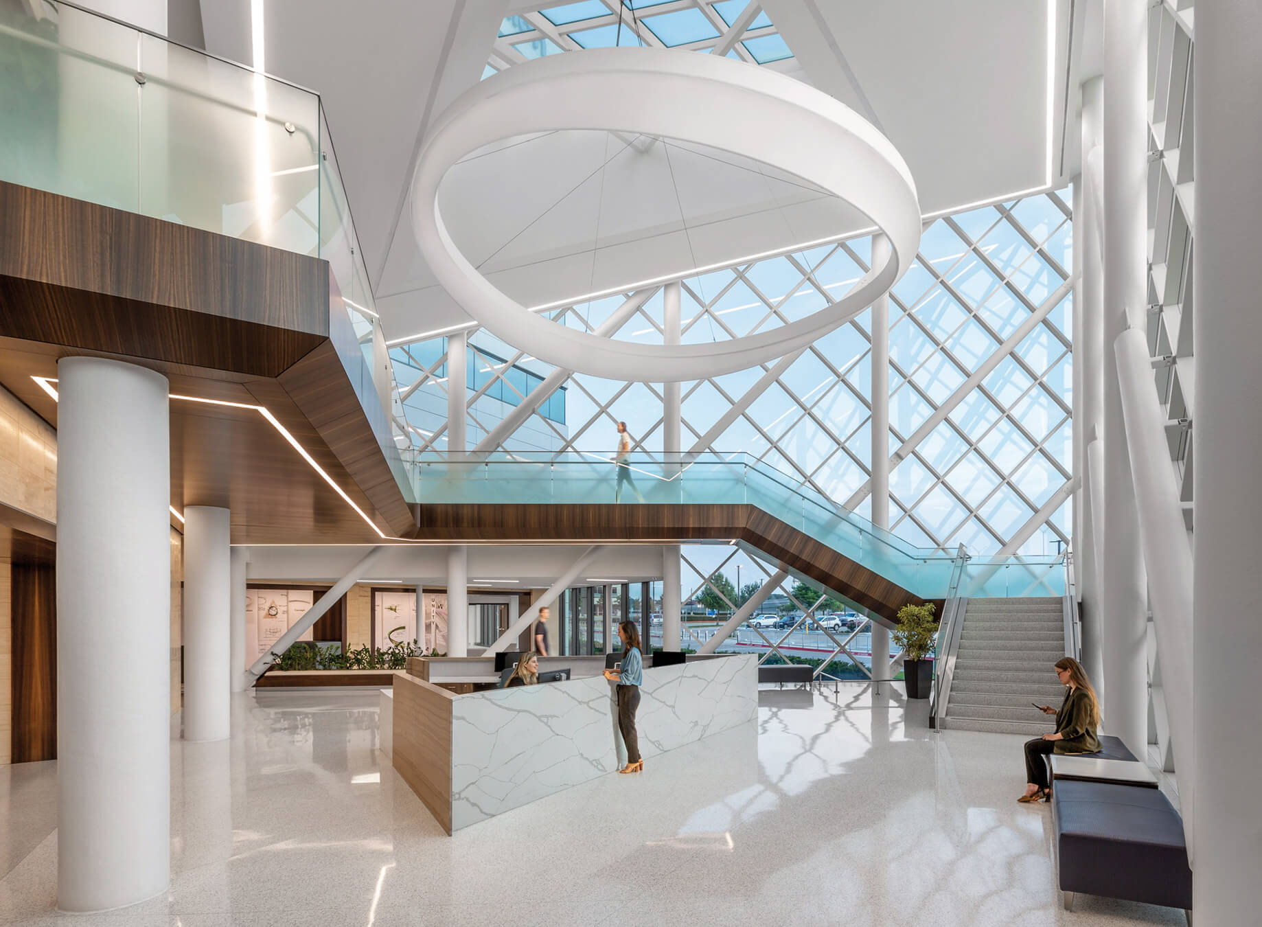 Interior view of a spacious hospital reception area featuring large glass windows, a circular light fixture, and modern staircases with people engaged in various activities.