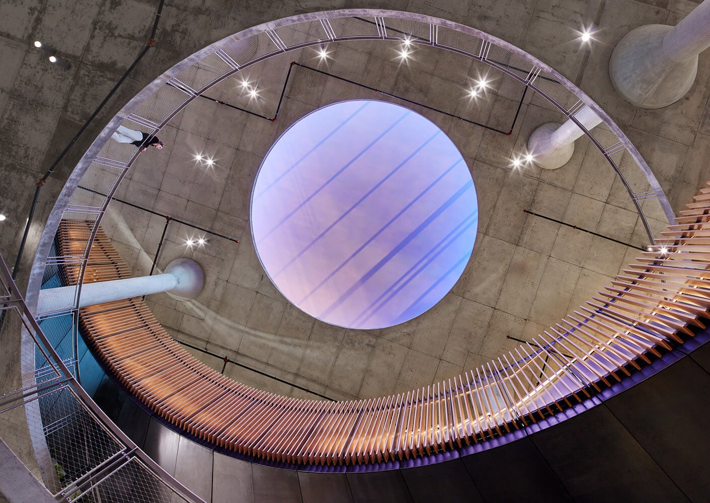 Aerial view of a circular architectural space featuring a lightwell, wooden seating, and concrete elements, illuminated by multiple ceiling lights.