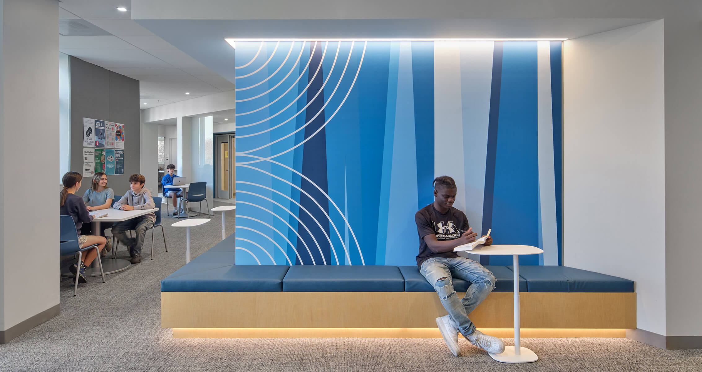 A student reads on a bench beside a blue-striped wall in a spacious area with seating, while others collaborate at a table nearby.