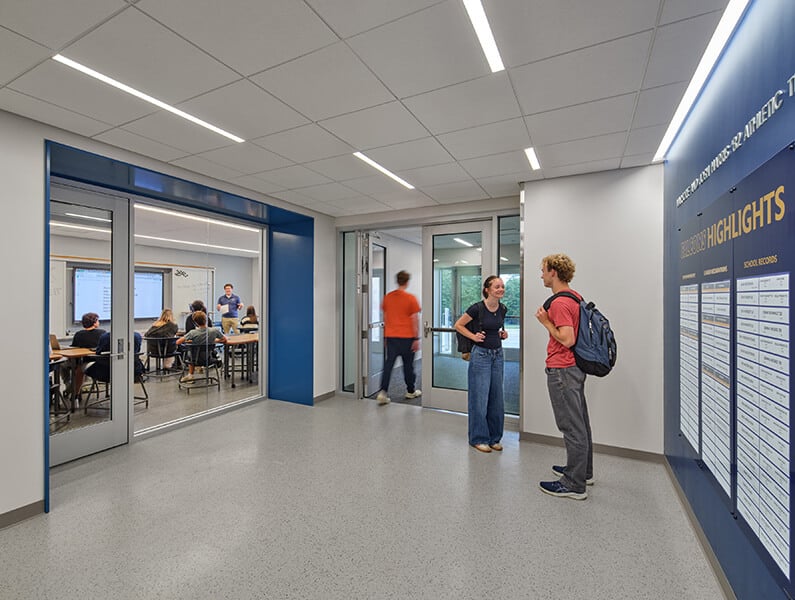 Two students converse in a hallway with large glass doors leading to a classroom. A whiteboard is visible in the background.