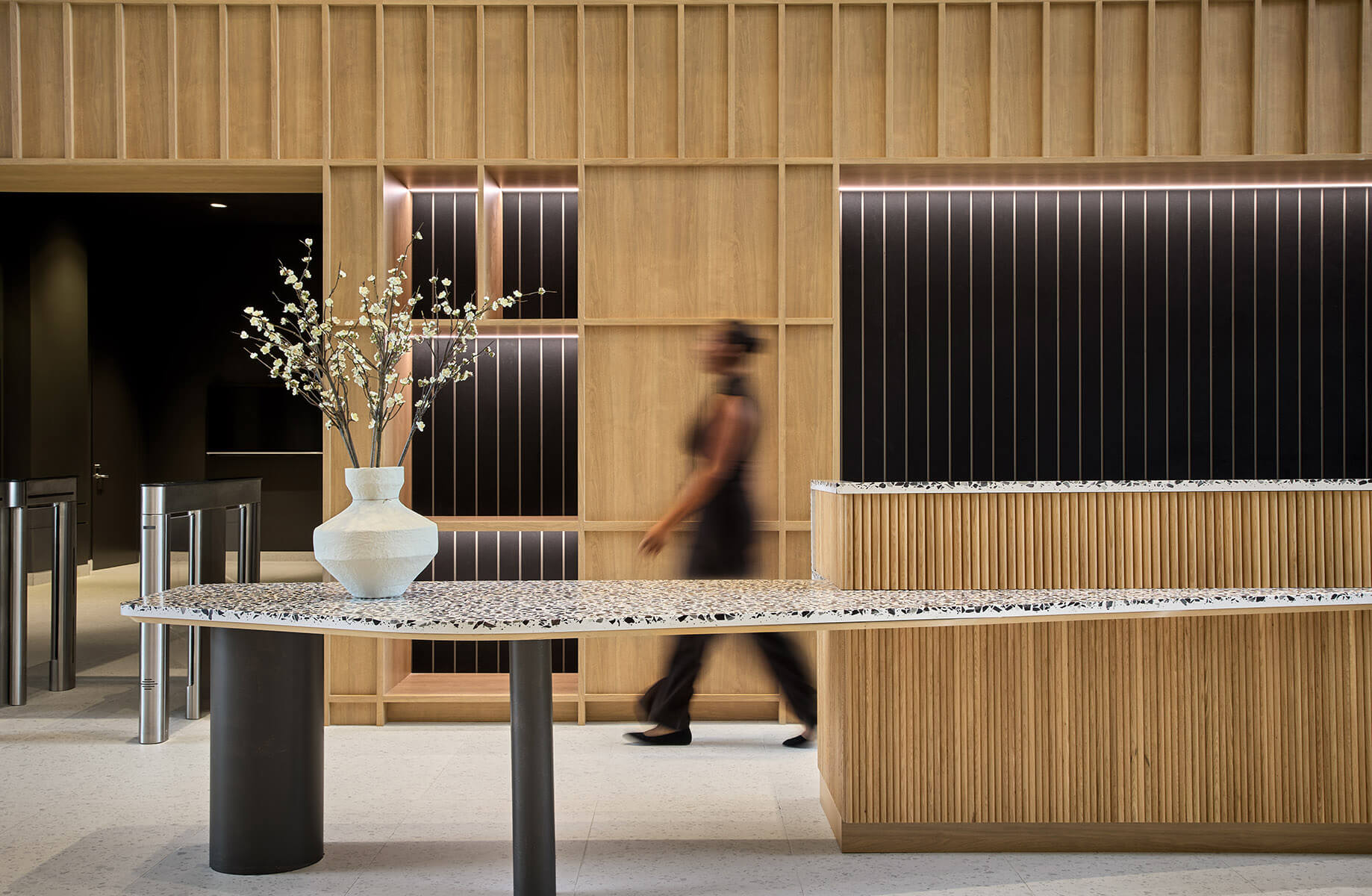 Reception area with a terrazzo counter, wooden paneling, and decorative vase. A blurred figure walks past, emphasizing a busy environment.