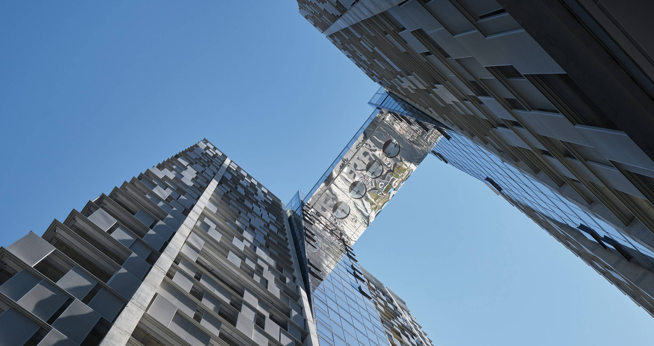 View of two tall buildings with textured facades, connected by a glass bridge against a clear blue sky.