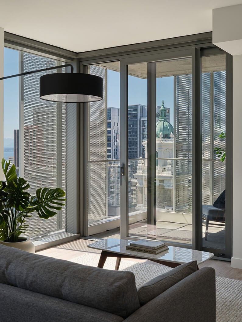 Interior view of a living space featuring a sofa, coffee table, potted plant, large windows with cityscape, and modern light fixture.