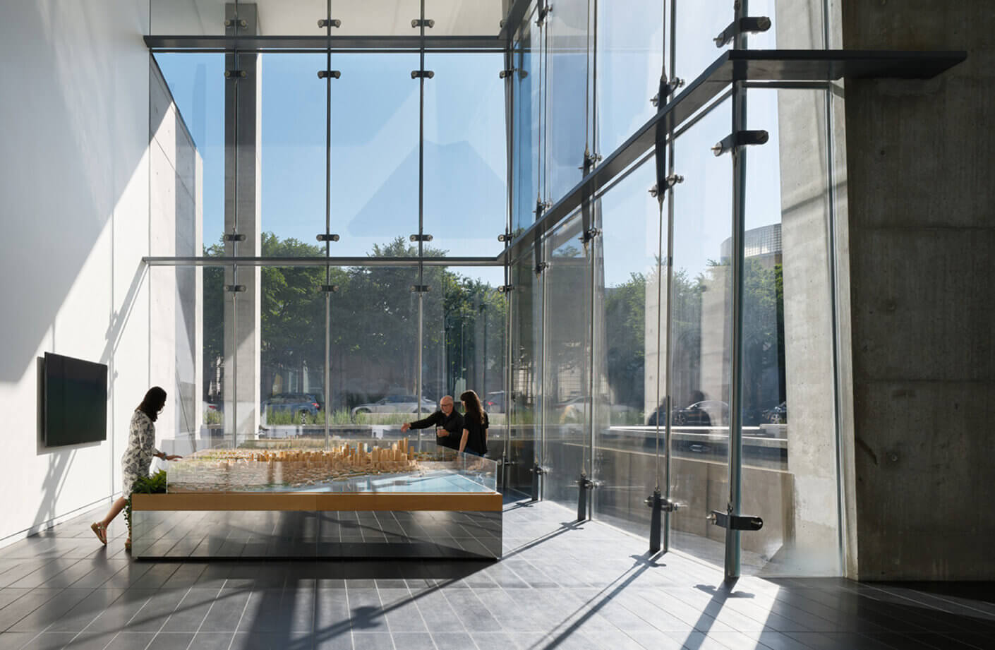 Two individuals engage with a scale model in a space featuring glass walls and concrete accents, with natural light illuminating the area.