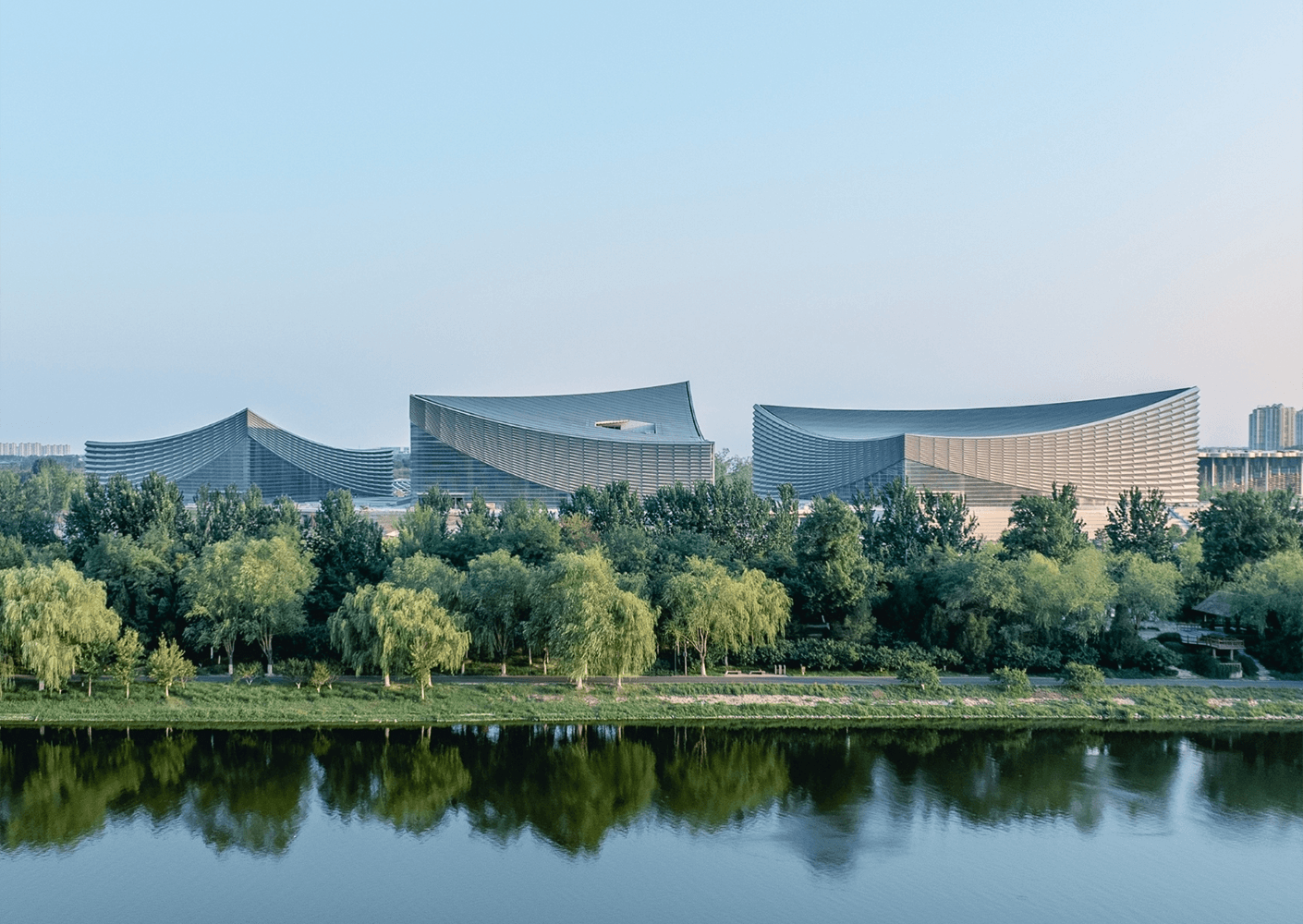 Three unique architectural structures with wavy roofs, surrounded by trees and reflecting in a river under a clear sky.