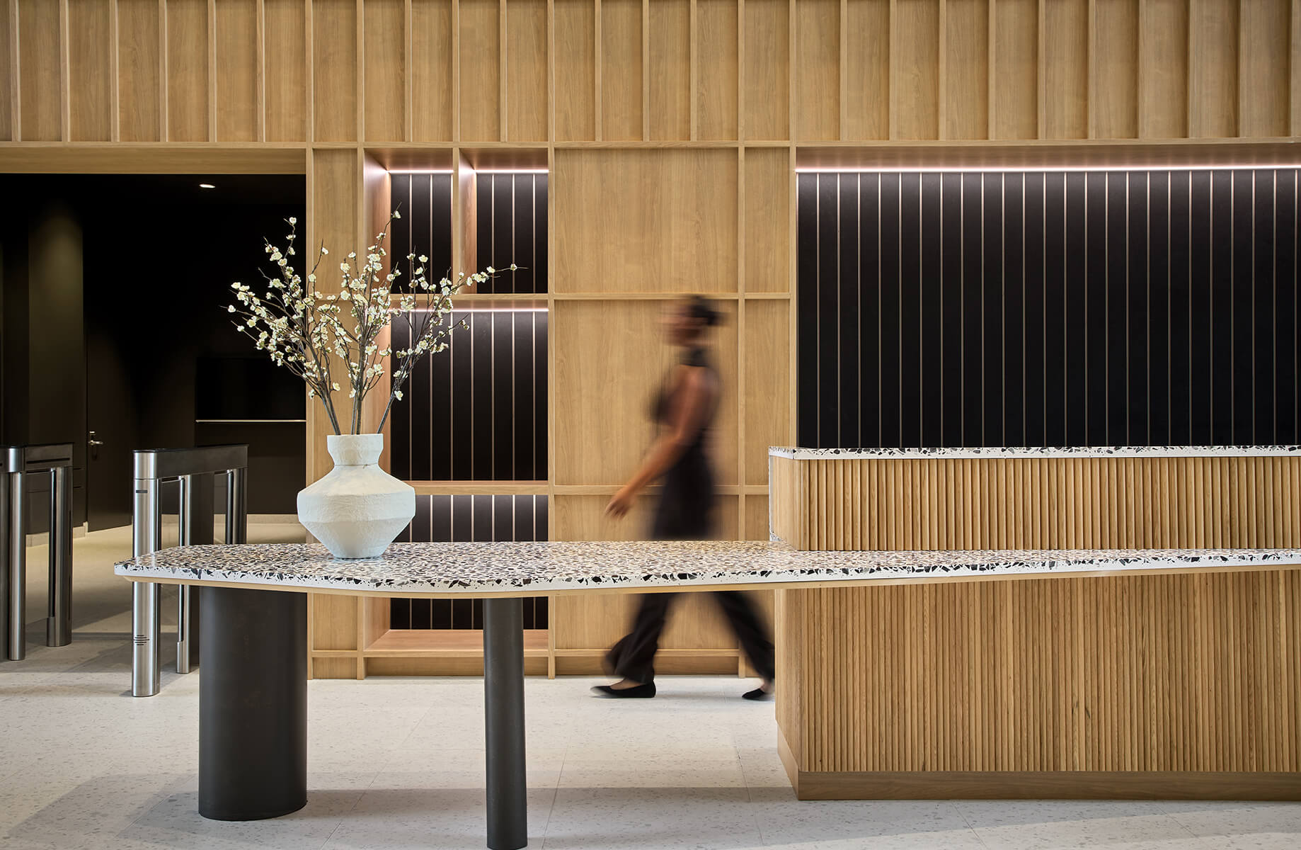 Reception area featuring a sleek desk with a textured surface, wooden accents, and a floral arrangement, with a person walking past.