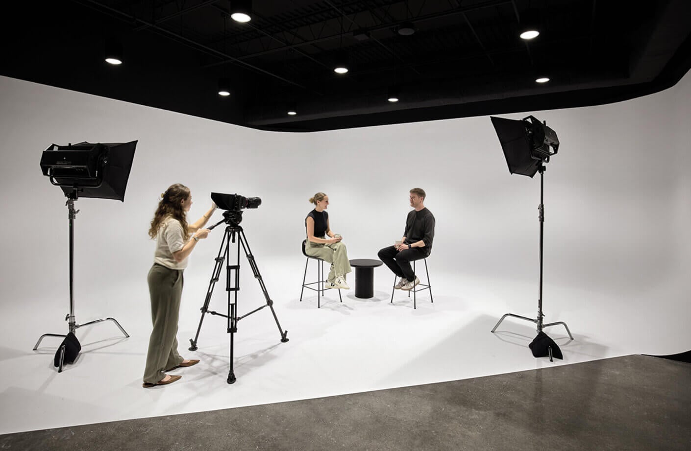 Two people seated for an interview on a minimalist set with a camera and lighting equipment in the background.