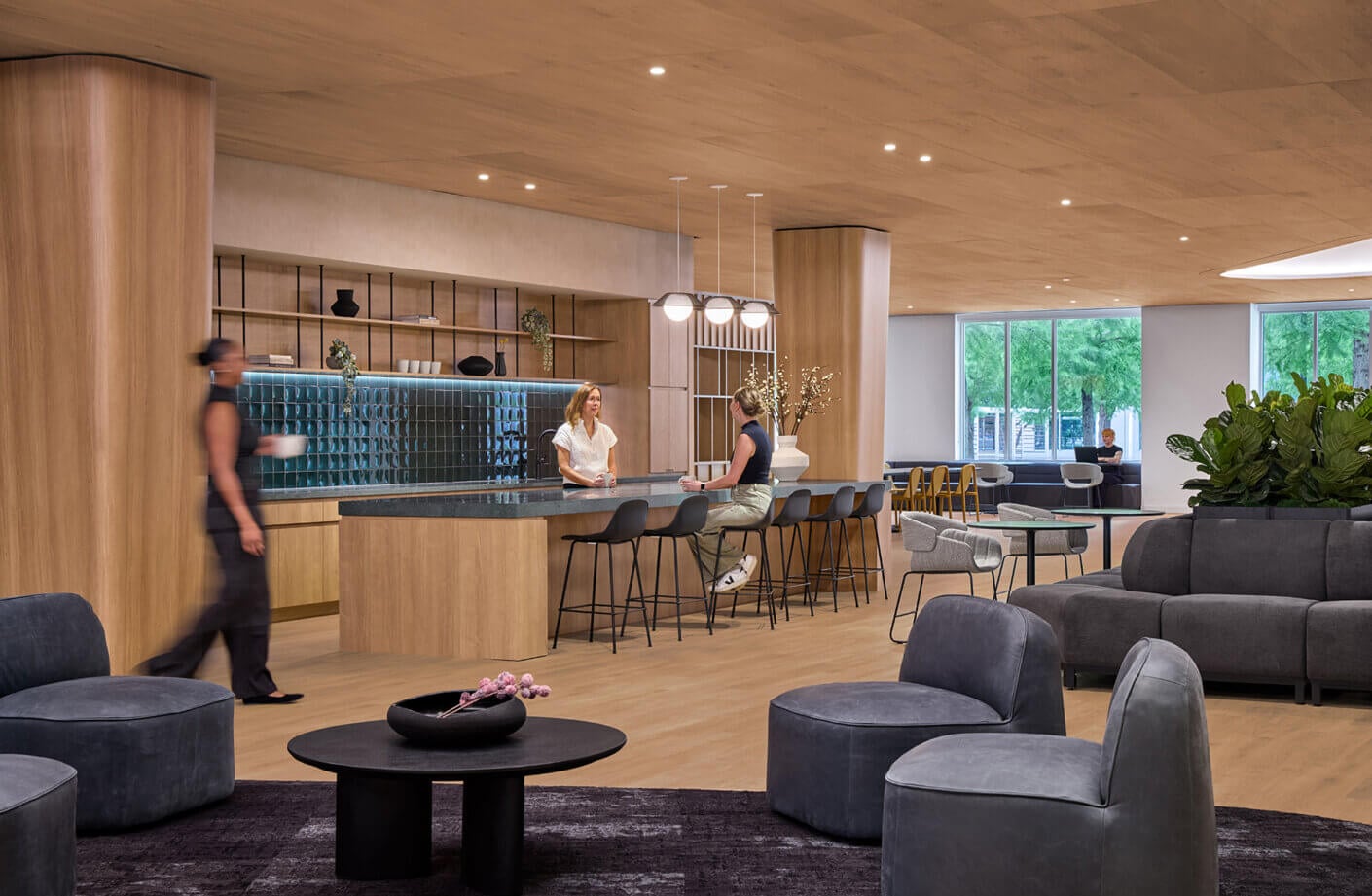 Two women converse at a sleek bar while a person walks past. The space features wood accents, comfortable seating, and modern decor elements.