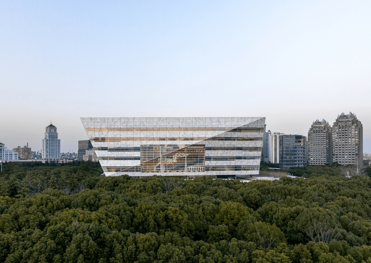 Glass building surrounded by lush greenery, with urban skyline in the background. Architectural design features sharp angles and reflections.