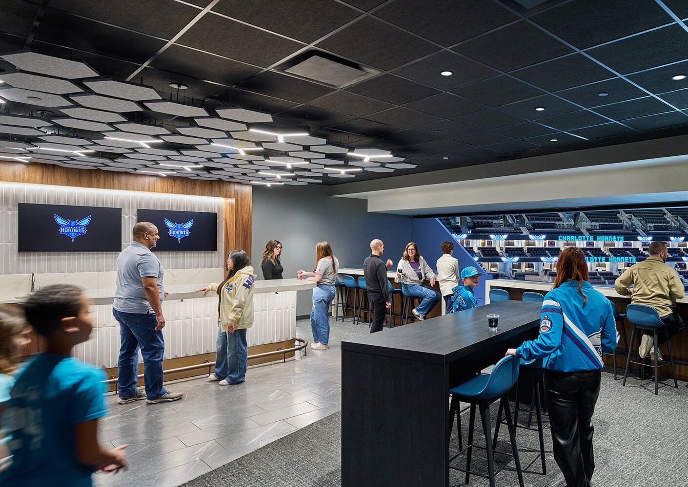 Group of people socializing in a stylish lounge area featuring the Charlotte Hornets logo, modern lighting, and a bar setting.