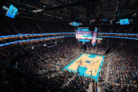 Aerial view of a packed Spectrum Center during a basketball game, featuring players on the court and a large video screen.